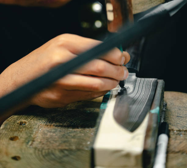 Close-up view of a skilled craftsman engraving intricate details onto the blade of a Japanese knife. The hands, steady and precise, guide the tool across the patterned steel, showcasing the care and mastery that goes into each piece. The reflection of light on the blade emphasizes its sharpness and fine craftsmanship.